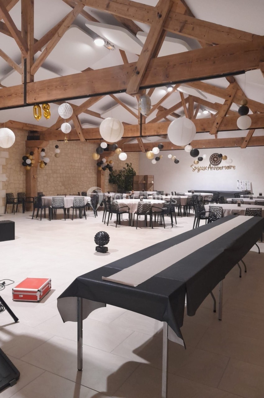 Salle décorée pour un anniversaire, avec tables alignées, guirlandes et lanternes suspendues sous une charpente en bois.