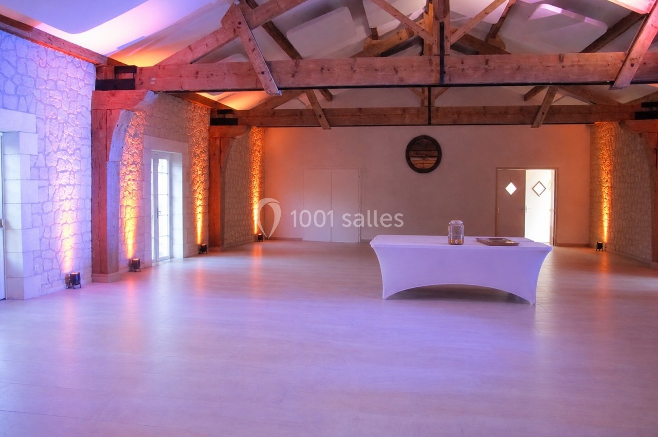 Grande salle lumineuse avec poutres apparentes, murs en pierre, éclairage chaleureux et table blanche au centre.