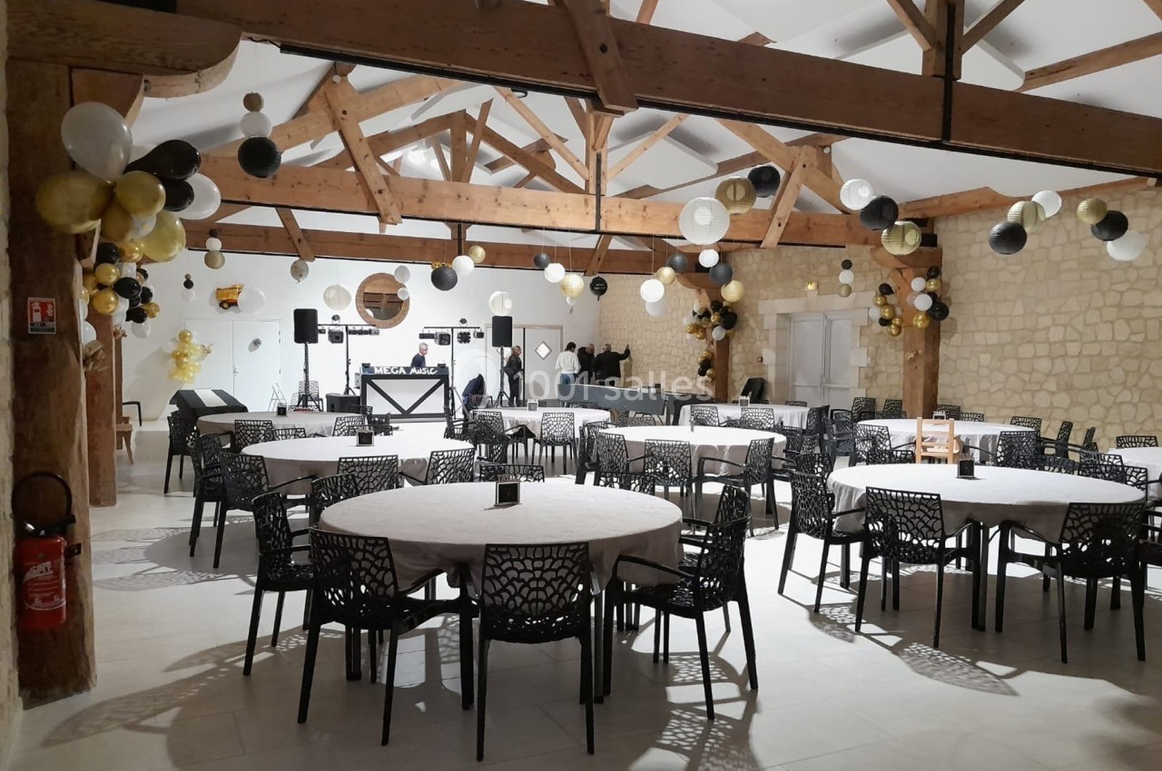 Salle de réception décorée avec des ballons noirs, blancs et dorés, tables rondes dressées et espace DJ au fond.