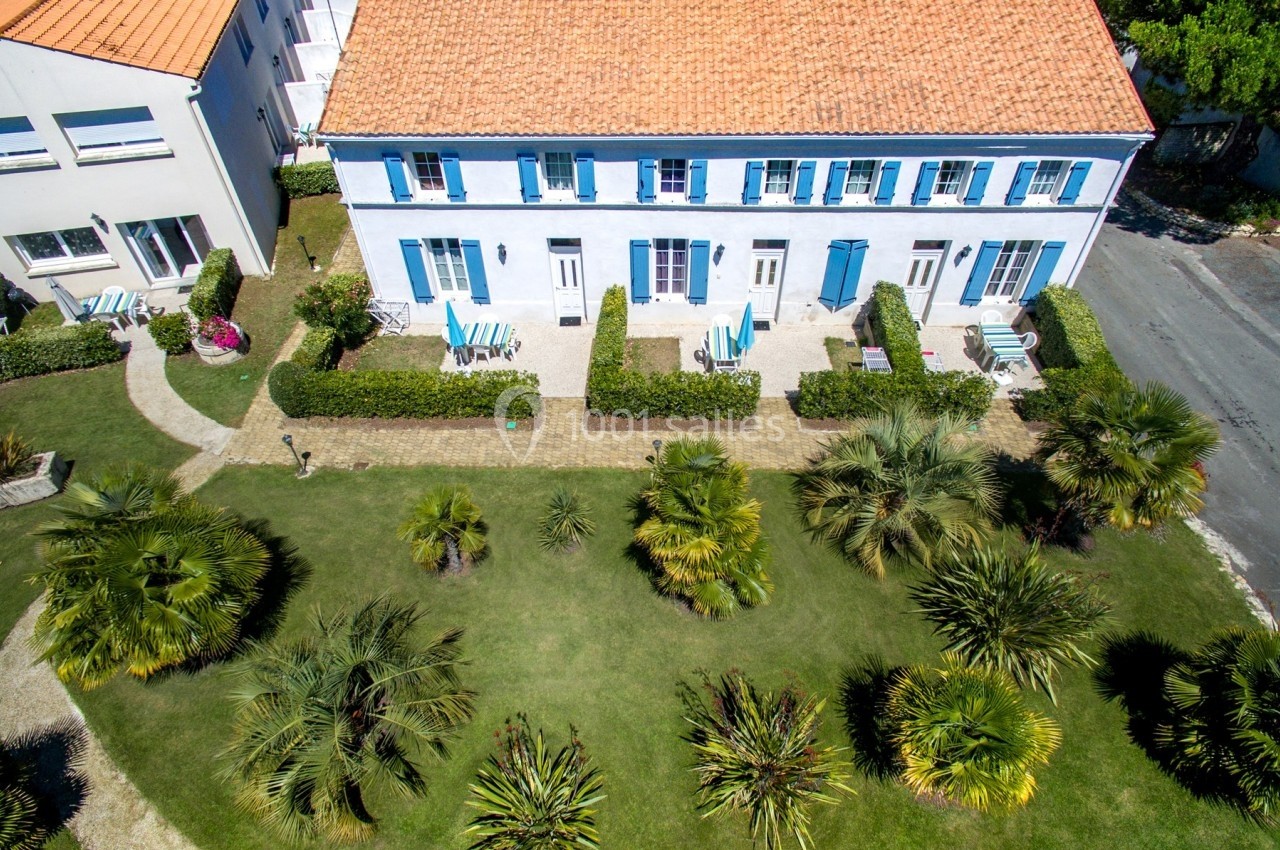 Vue aérienne d'une maison à deux étages avec façade blanche et volets bleus, entourée d'un jardin arboré.