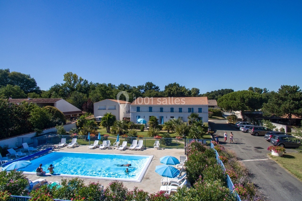 Vue aérienne d'une piscine extérieure entourée de transats, avec un bâtiment blanc et un parking en arrière-plan.