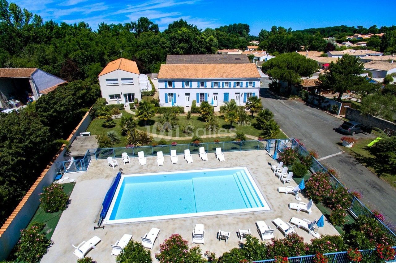 Vue aérienne d'une piscine extérieure entourée de transats, avec un bâtiment blanc et un jardin en arrière-plan.