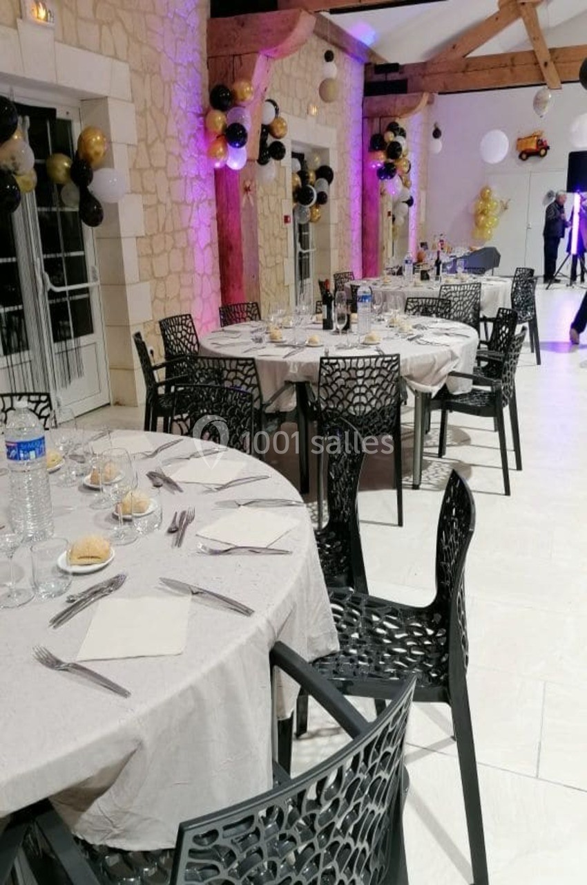 Salle de réception décorée avec des ballons, tables dressées avec nappes blanches et chaises noires ajourées.
