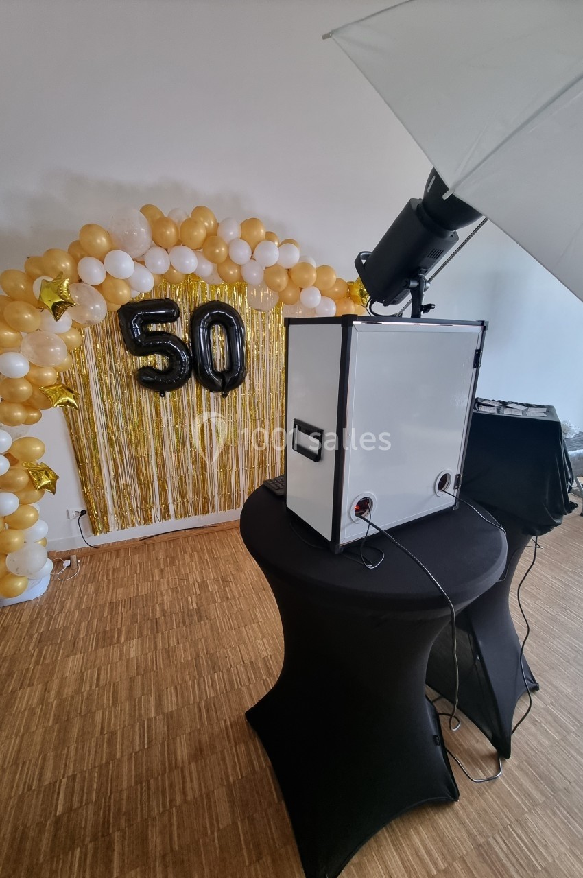 Décoration festive avec arche de ballons dorés et blancs, chiffre 50 en ballons noirs, et équipement photo en avant-plan.