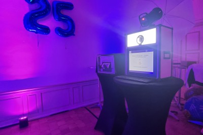 Borne photo rétro-éclairée dans une salle décorée avec des ballons ’25’ et un éclairage violet.