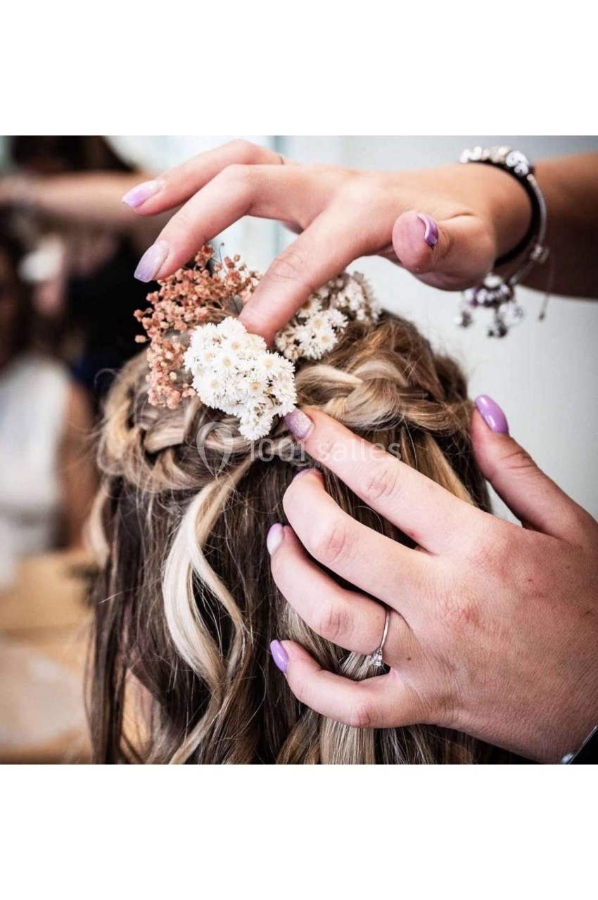Coiffure tressée ornée de fleurs blanches et roses, ajustée par des mains féminines avec des ongles vernis.