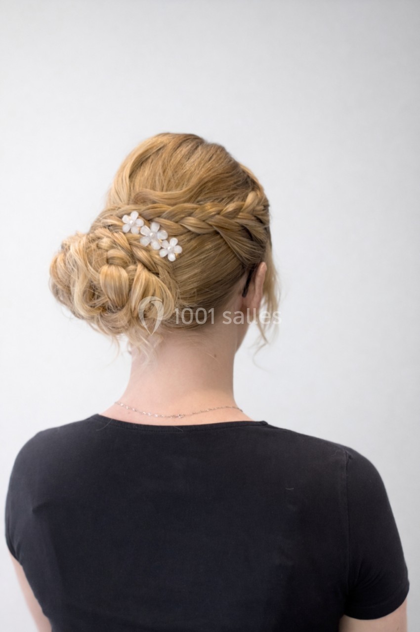 Coiffure blonde avec tresses et chignon, ornée d'une barrette à fleurs blanches, vue de dos.