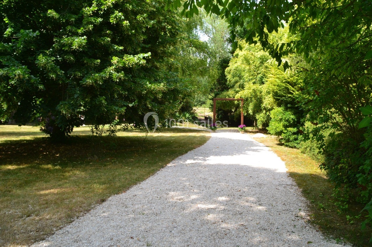Allée de gravier bordée d'arbres et de verdure, menant à une arche fleurie dans un jardin.