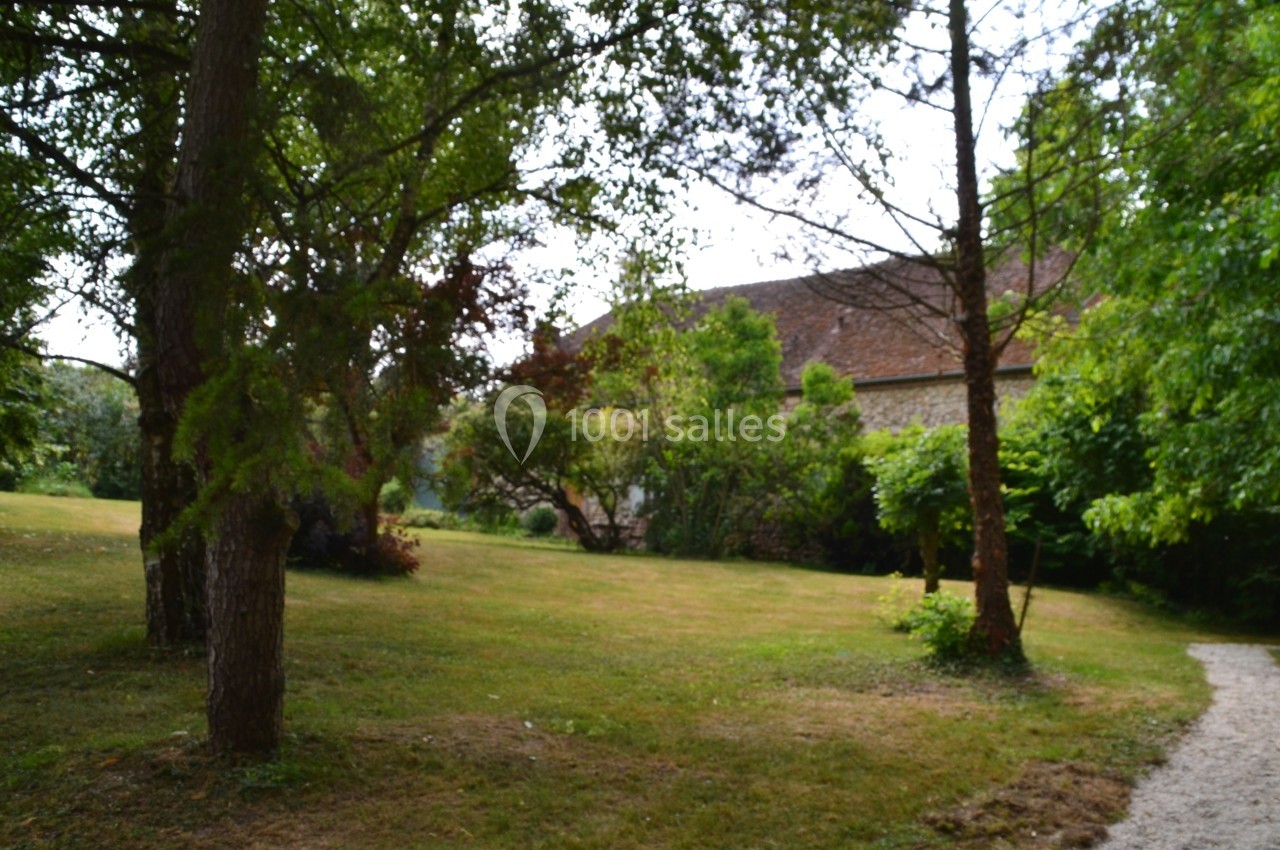 Jardin verdoyant avec pelouse, arbres et arbustes, bordé par un chemin en gravier et une maison en pierre au fond.