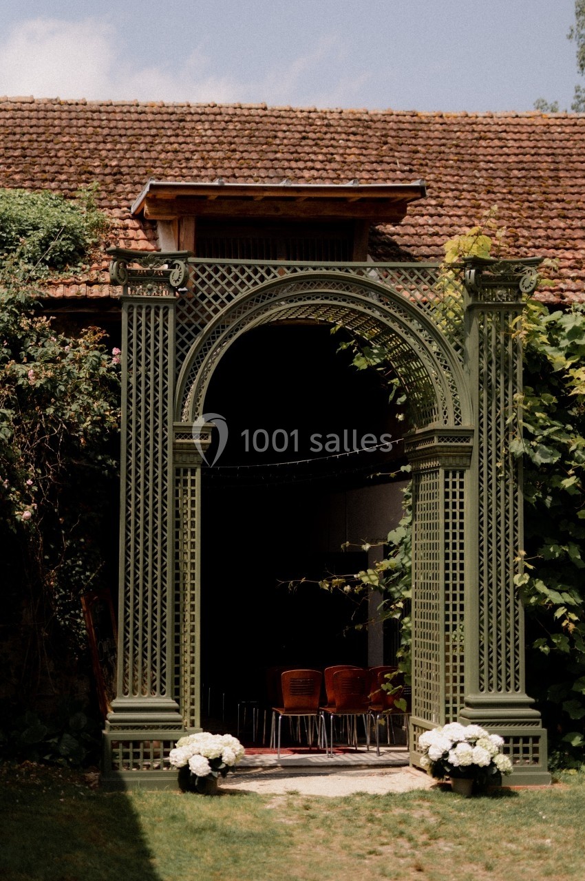 Arche en bois vert ornée de plantes, donnant sur un espace intérieur avec des chaises alignées.