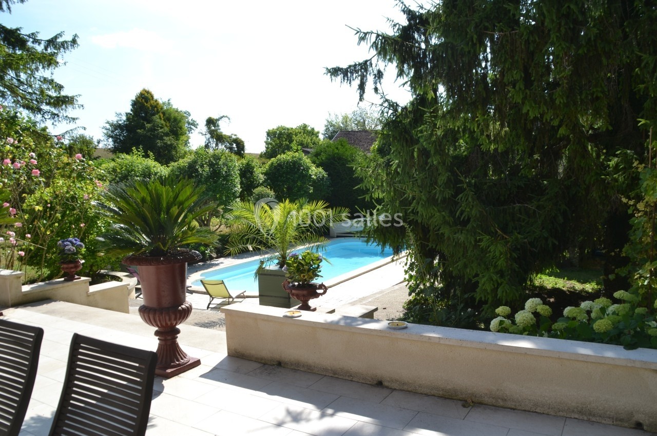 Terrasse avec chaises donnant sur un jardin verdoyant et une piscine entourée de plantes et d'arbres.
