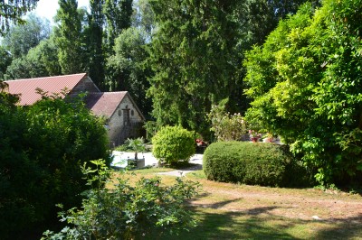 Jardin verdoyant avec pelouse, arbres et arbustes, bordé par un chemin en gravier et une maison en pierre au fond.