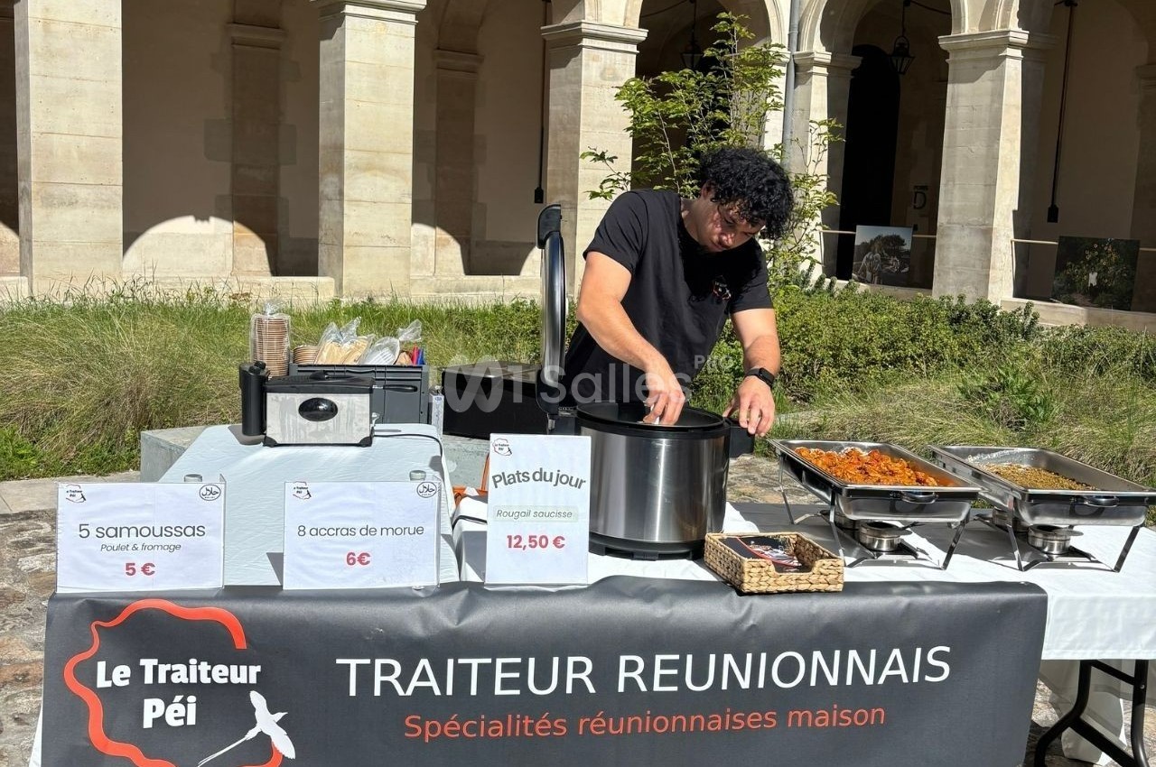 Un traiteur prépare des plats réunionnais derrière un stand avec menus affichés, dans une cour ensoleillée.