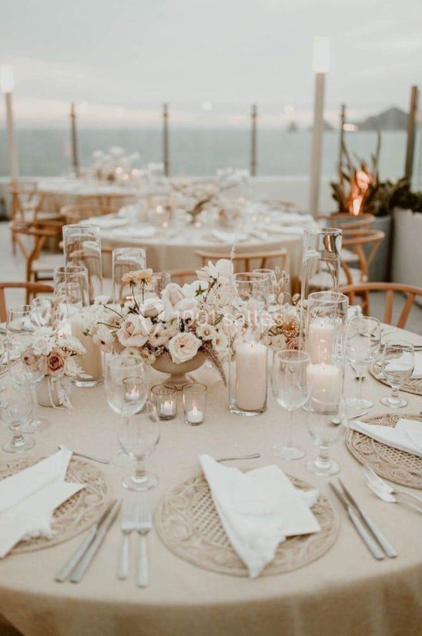 Tables élégamment dressées avec nappes beiges, fleurs blanches, bougies et vue sur la mer au crépuscule.