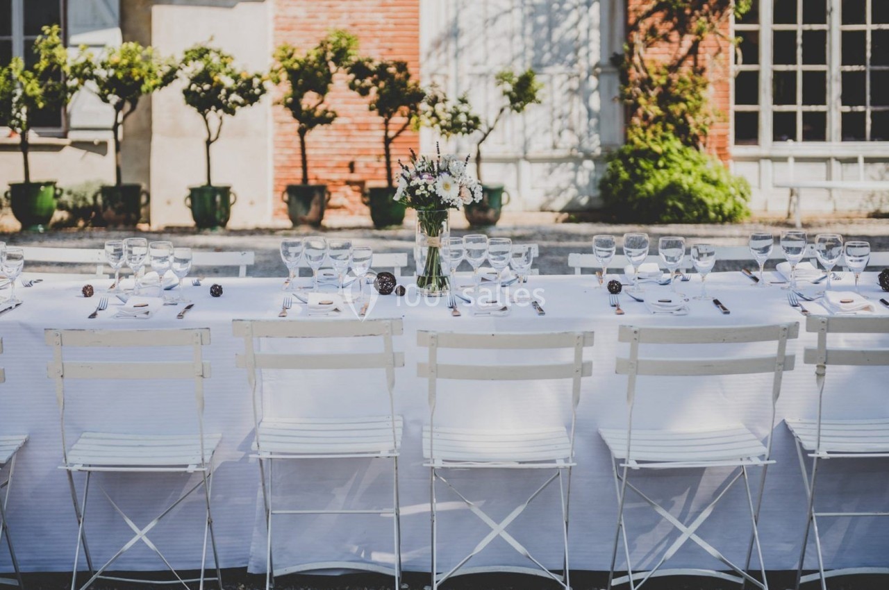 Table dressée en extérieur avec nappes blanches, chaises pliantes et centre de table floral, devant un bâtiment en briques.