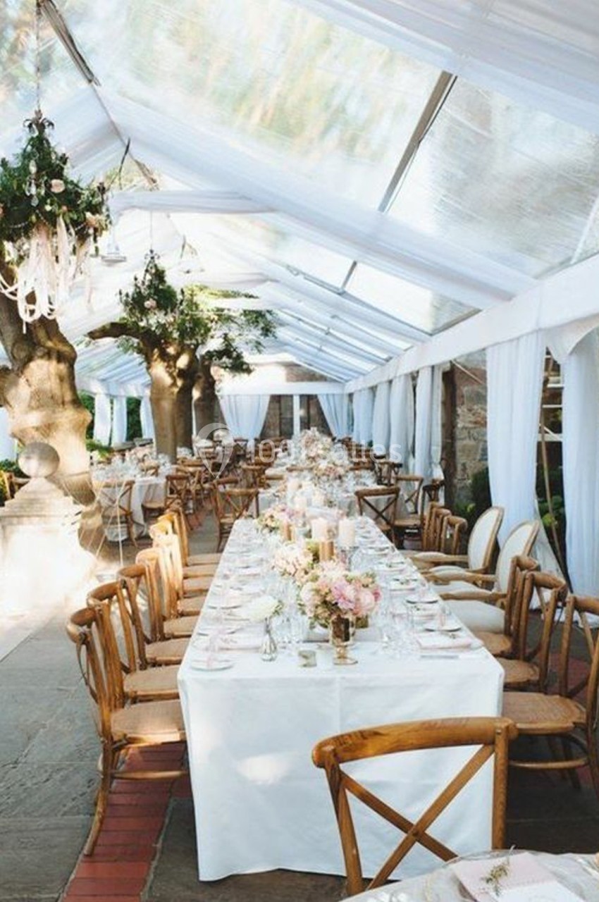 Tables décorées avec des fleurs et nappes blanches sous une tente transparente, entourées de chaises en bois.