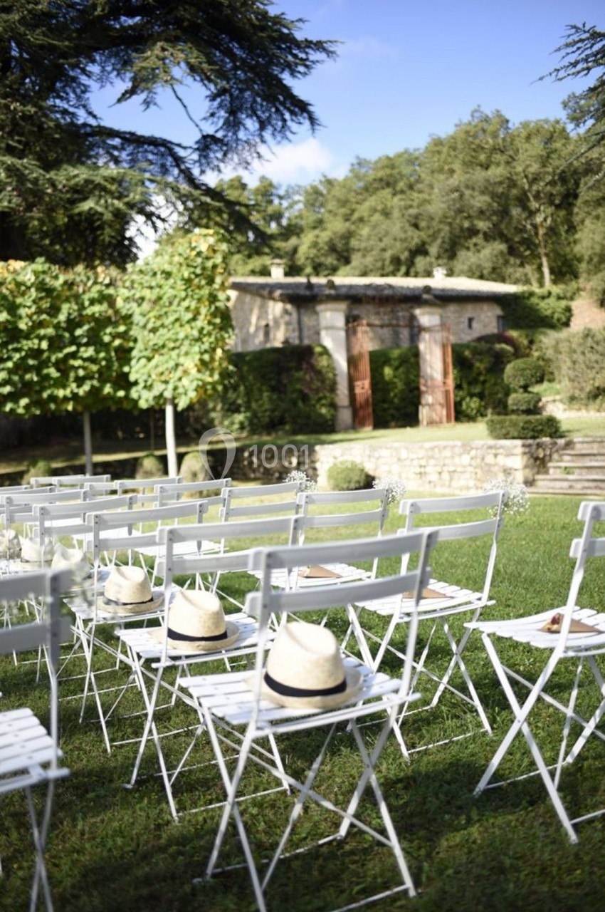 Chaises blanches alignées sur une pelouse, avec des chapeaux posés dessus, devant un jardin et une bâtisse en pierre.