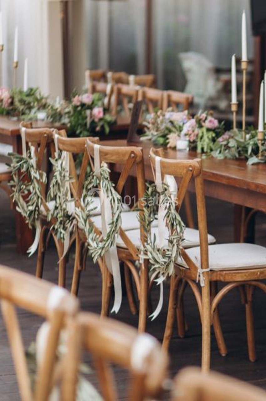 Chaises en bois décorées de couronnes de feuillage et rubans, alignées autour d'une table ornée de fleurs et bougies.