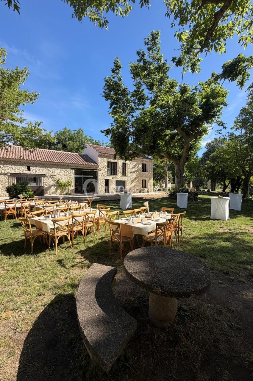 Tables dressées pour un événement en plein air dans un jardin ombragé, avec une maison en pierre en arrière-plan.