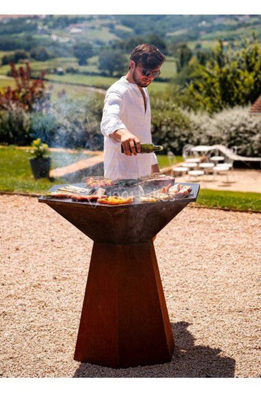 Homme en chemise blanche cuisant des aliments sur un brasero dans un jardin avec vue sur la campagne.