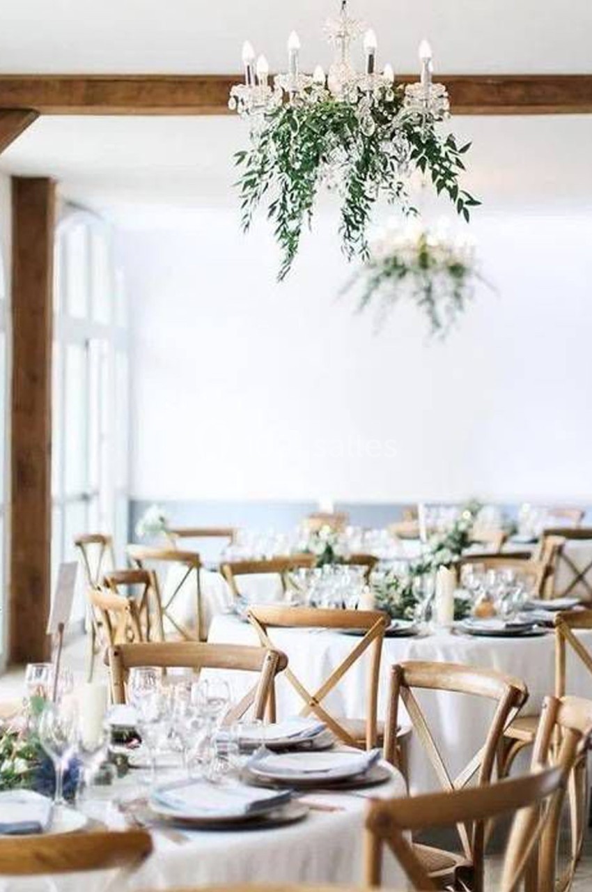 Salle de réception lumineuse avec tables décorées, chaises en bois et lustres ornés de feuillage suspendu.