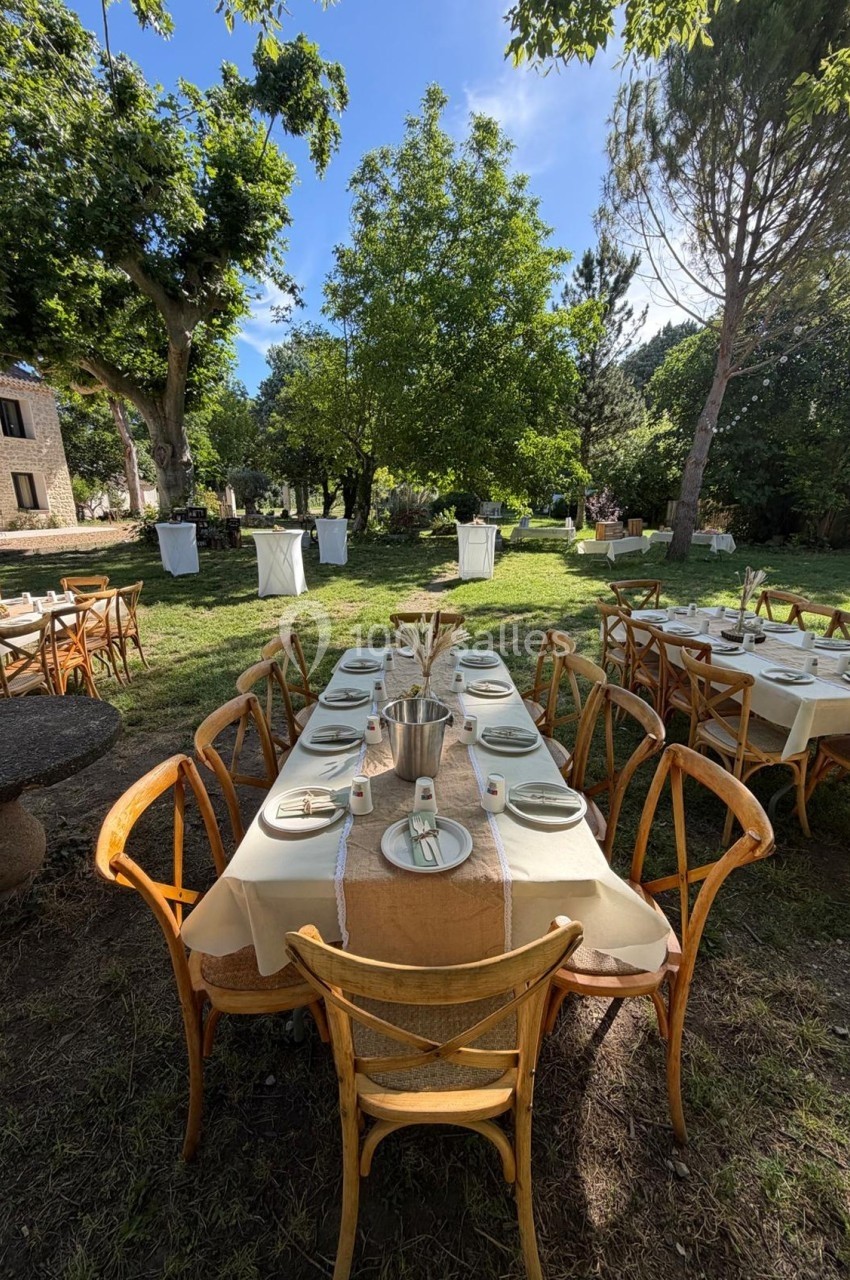 Tables en bois dressées avec nappes et couverts, disposées en extérieur dans un jardin arboré par temps ensoleillé.
