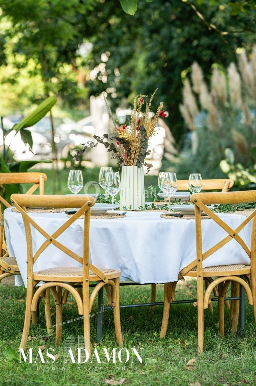 Table ronde dressée en extérieur avec nappe blanche, chaises en bois et vase de fleurs séchées au centre.