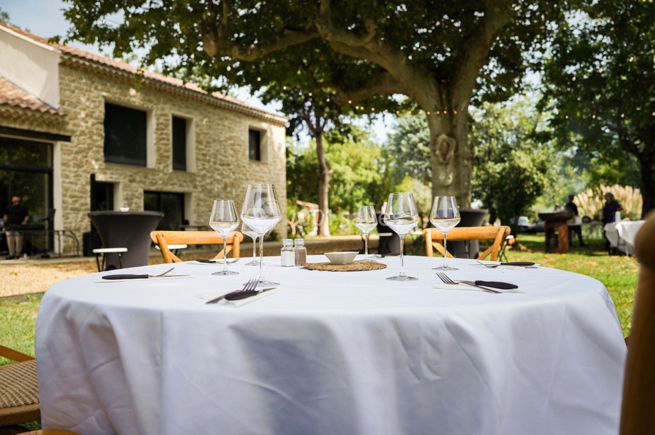 Table ronde dressée avec nappe blanche, verres à vin et couverts, en extérieur près d'une maison en pierre.