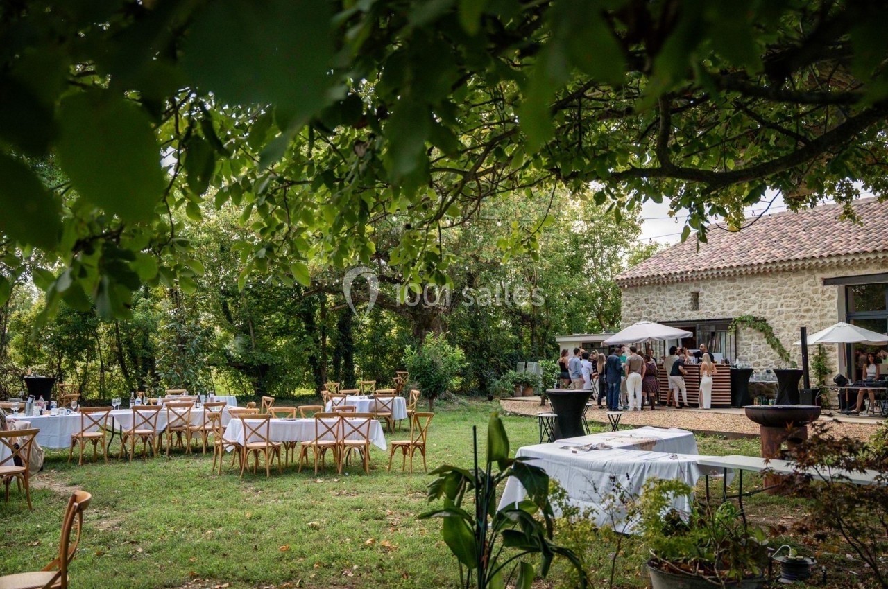 Tables dressées en plein air sous des arbres, près d'une bâtisse en pierre, avec des invités rassemblés au loin.