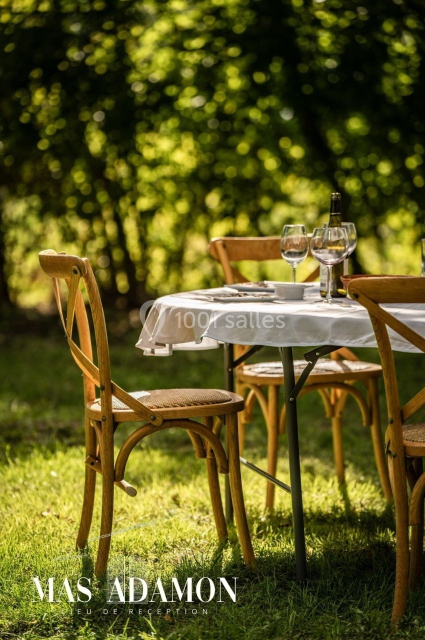 Table dressée en extérieur avec nappage blanc, chaises en bois, verres à vin et bouteille, sur fond de verdure.