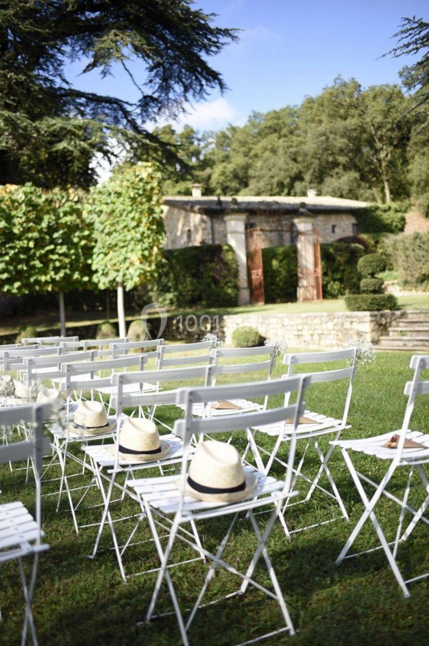 Chaises blanches alignées sur une pelouse, avec des chapeaux posés dessus, devant un jardin et une bâtisse en pierre.