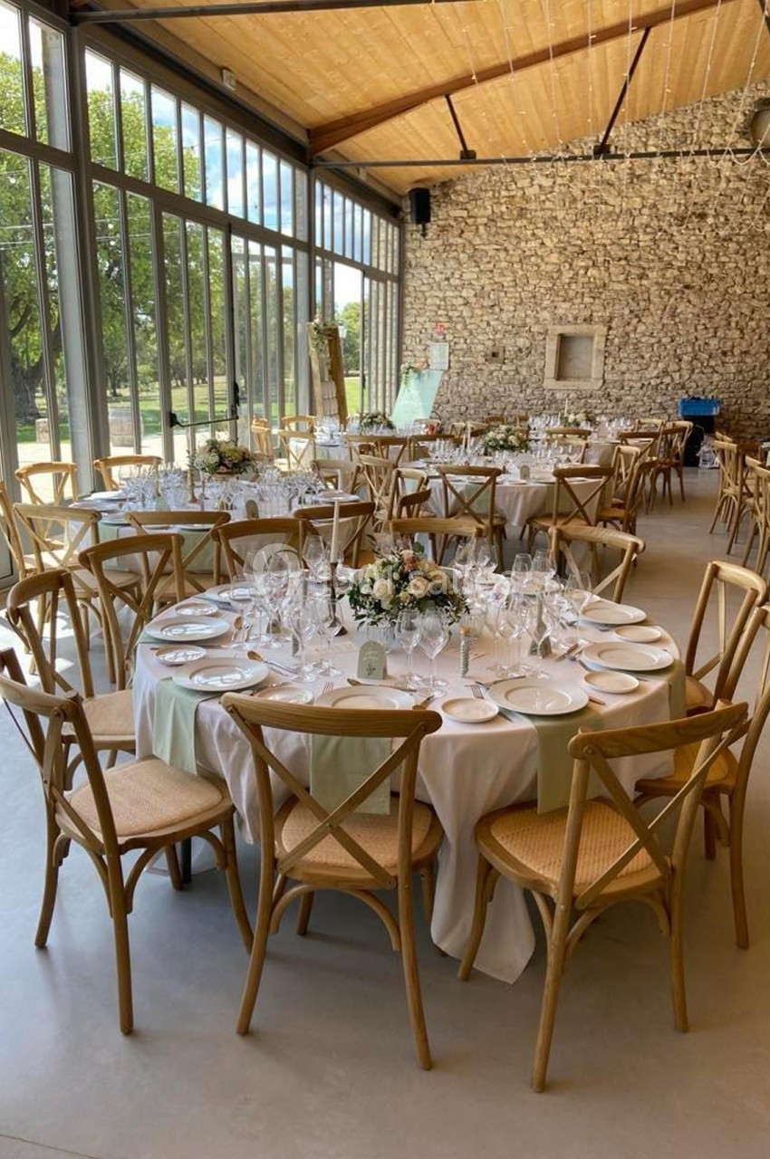 Salle lumineuse avec tables rondes décorées pour un événement, chaises en bois et murs en pierre apparente.
