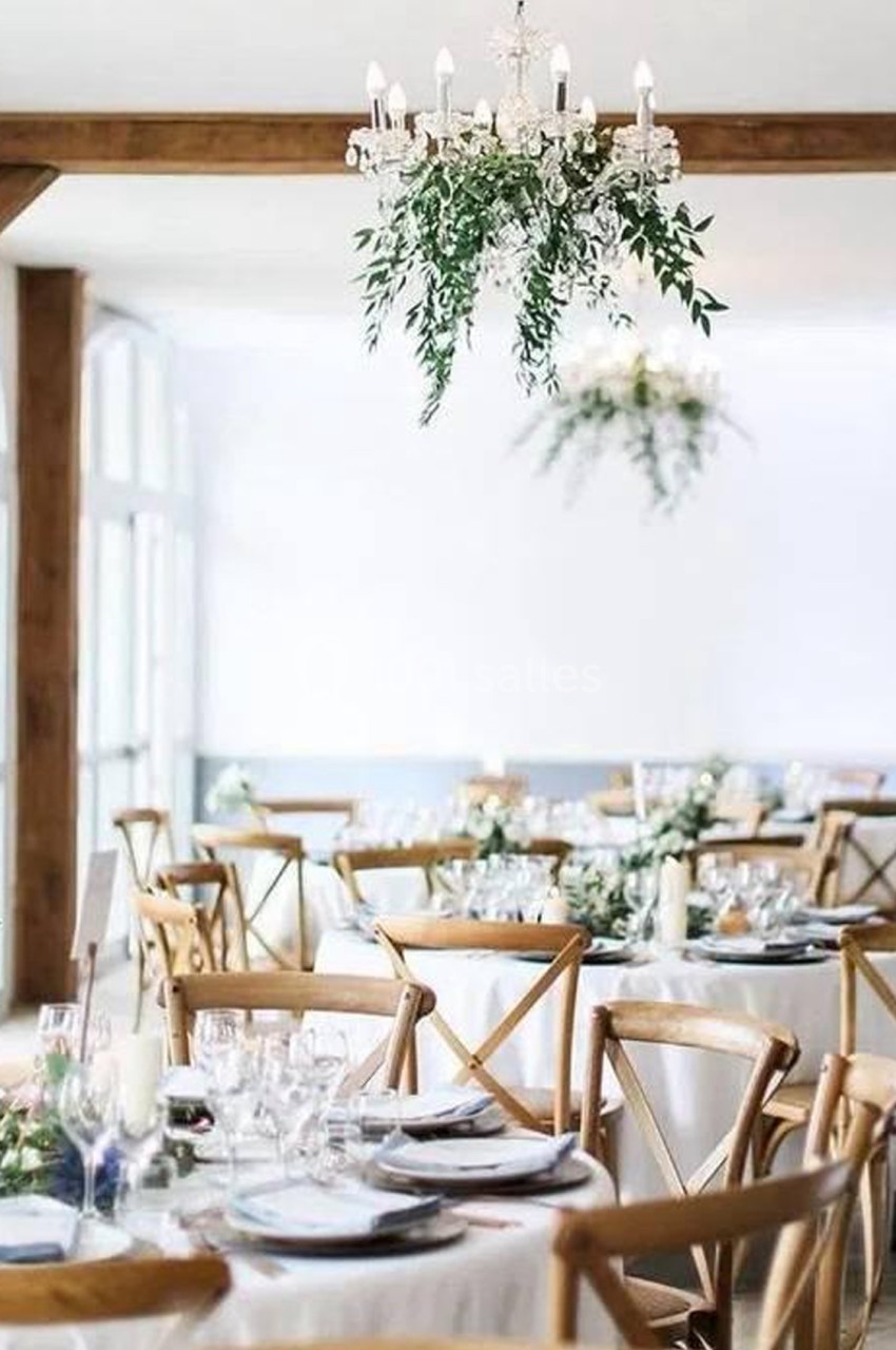 Salle de réception lumineuse avec tables rondes décorées, chaises en bois et lustres ornés de feuillage.
