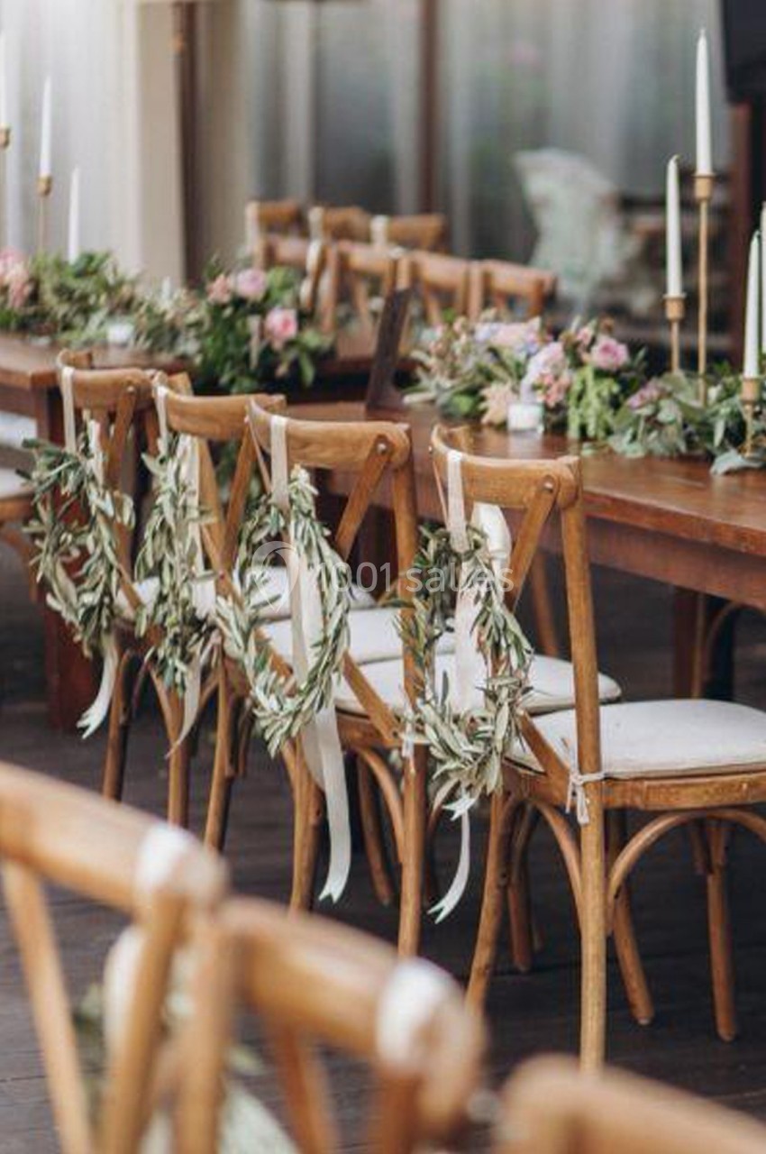 Chaises en bois décorées de couronnes de feuillage et rubans blancs autour d'une table ornée de fleurs et bougies.