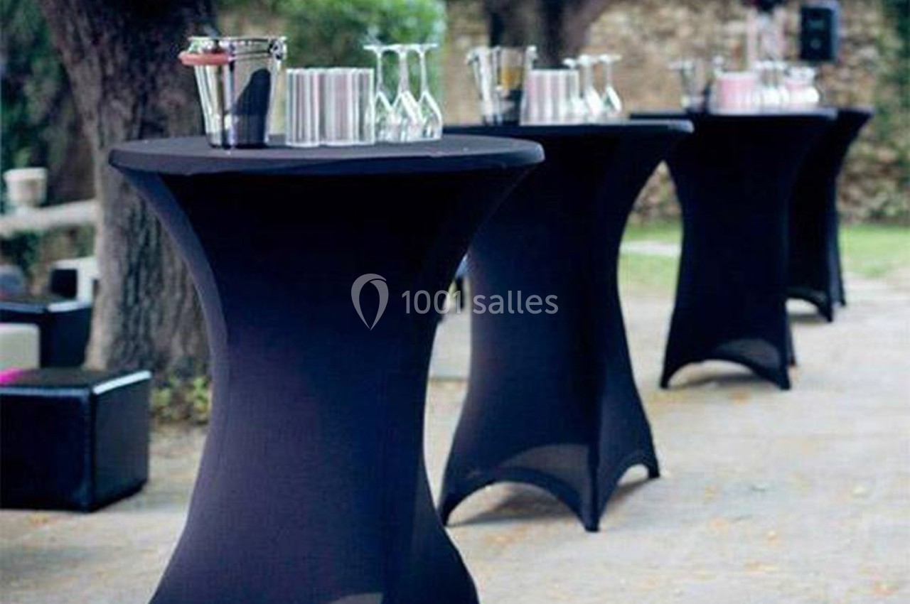 Tables hautes noires avec nappes, disposées en extérieur, garnies de verres et accessoires de bar.