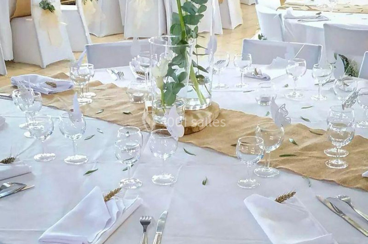 Salle de réception décorée pour un événement, avec tables rondes dressées, nappes blanches et éléments végétaux.