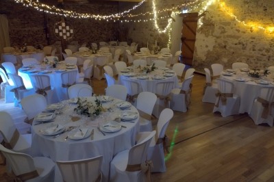 Salle de réception décorée avec des guirlandes lumineuses, tables rondes dressées et chaises blanches.