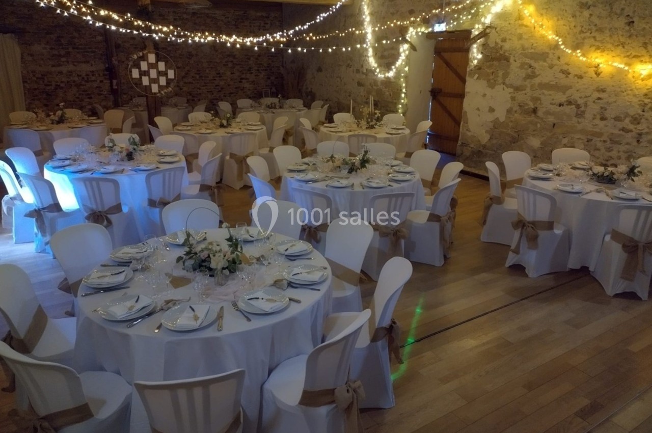 Salle de réception décorée pour un mariage, avec tables rondes dressées, chaises ornées et guirlandes lumineuses suspendues.