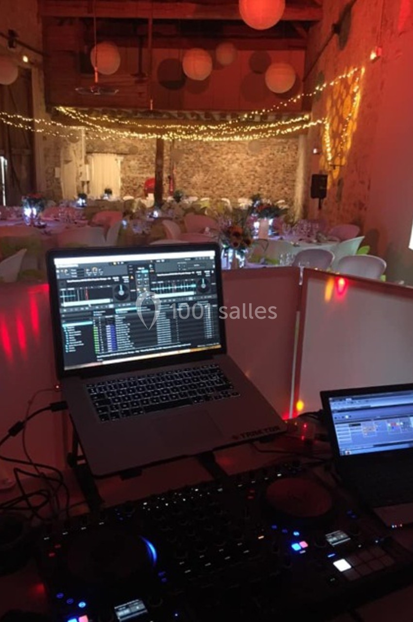 Poste de DJ avec ordinateurs et platines dans une salle décorée de guirlandes lumineuses et tables dressées pour un…