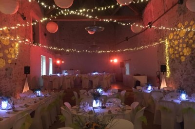Salle de réception décorée avec des guirlandes lumineuses, tables rondes dressées et chaises blanches.