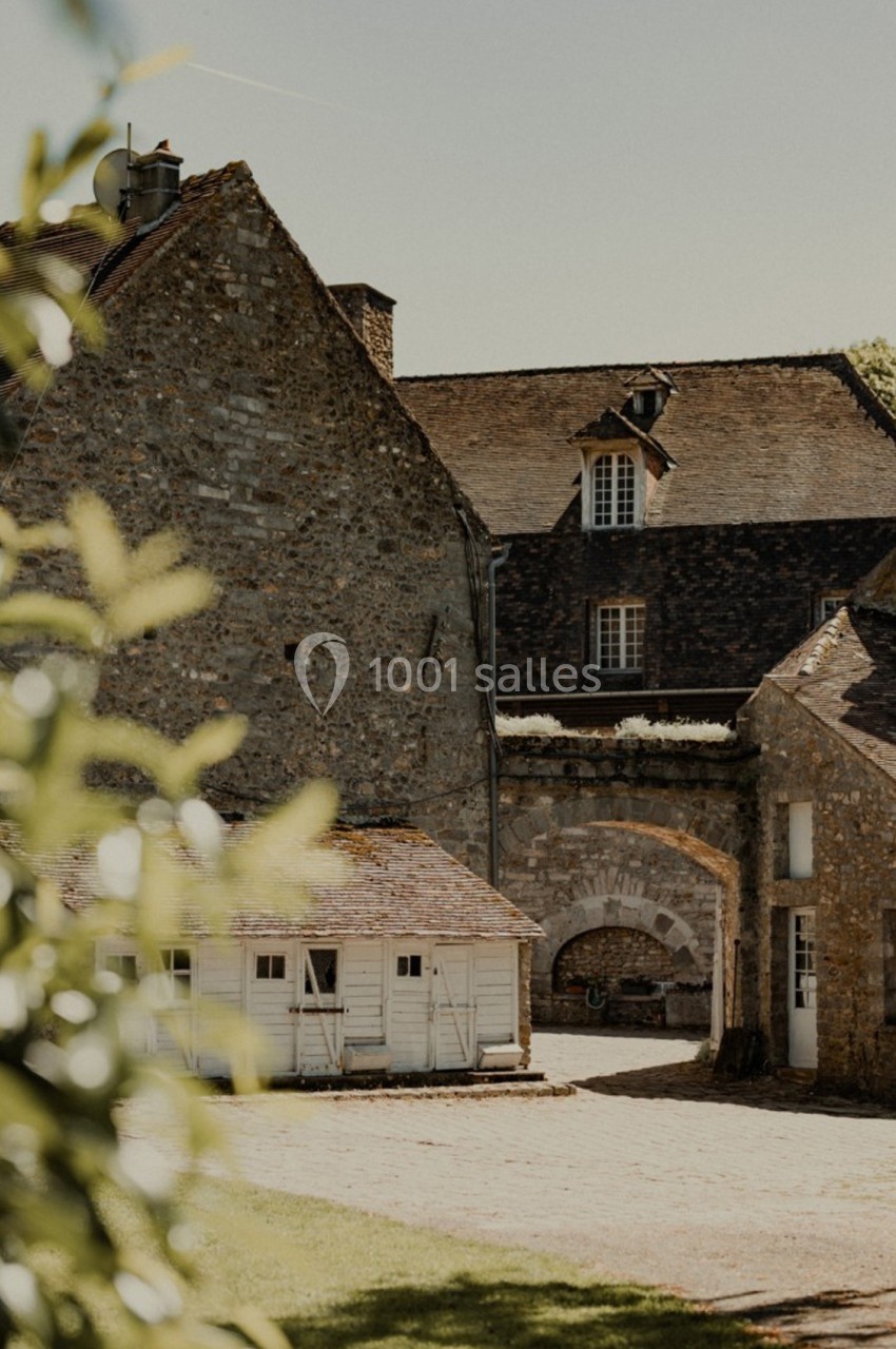 Bâtiments en pierre avec toits en ardoise entourant une cour, partiellement ombragés par des feuillages au premier plan.