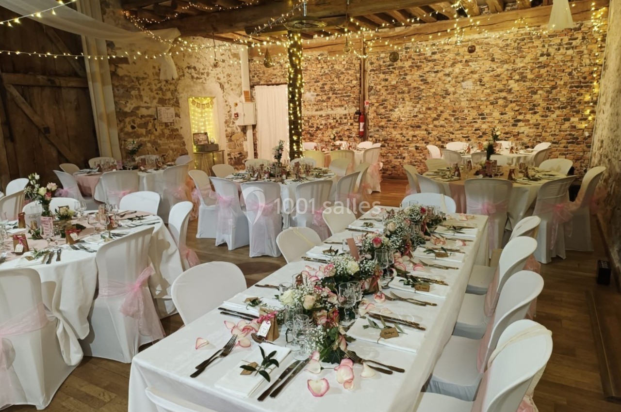 Salle de réception décorée pour un mariage, avec tables dressées, nappes blanches, fleurs et guirlandes lumineuses.