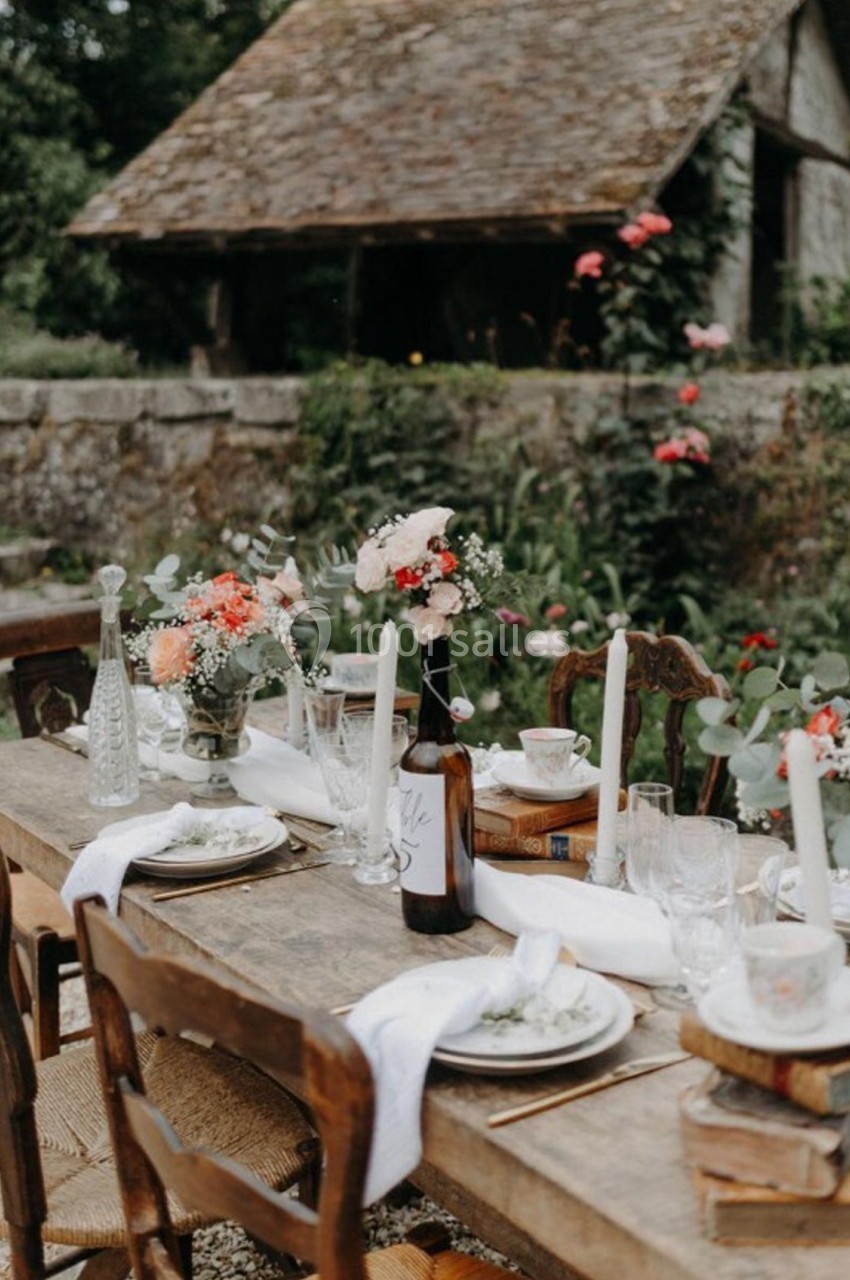 Table en bois rustique décorée de fleurs, bougies et vaisselle, installée dans un jardin près d'une maison en pierre.