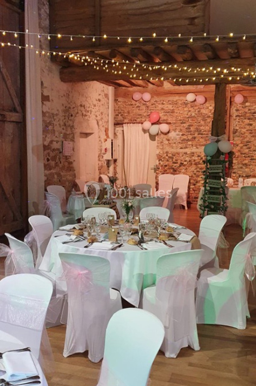 Salle de réception décorée pour un événement, avec tables rondes dressées, chaises blanches et guirlandes lumineuses.