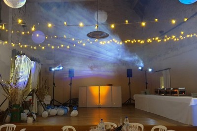 Salle de réception décorée avec des guirlandes lumineuses, tables rondes dressées et chaises blanches.