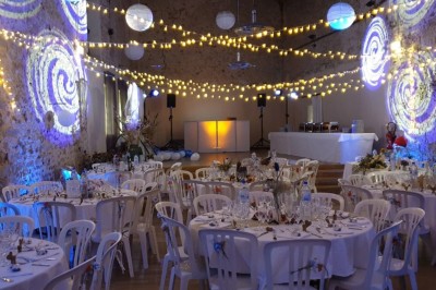 Salle de réception décorée avec des guirlandes lumineuses, tables rondes dressées et chaises blanches.