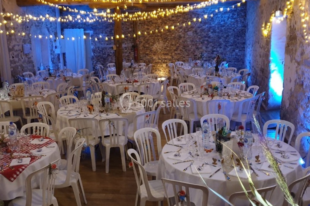 Salle de réception décorée avec des guirlandes lumineuses, tables rondes dressées et chaises blanches.