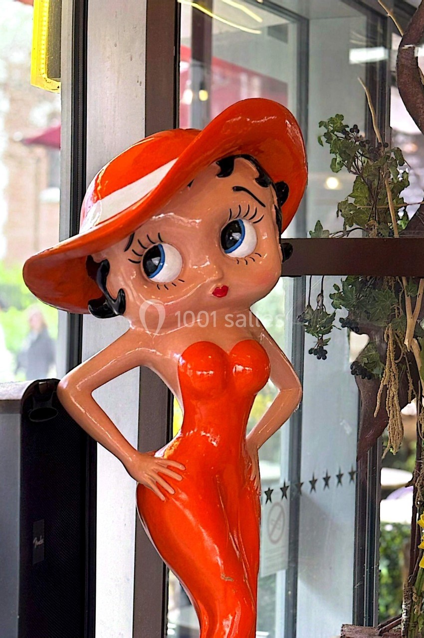 Statue colorée d'une femme stylisée en robe orange et chapeau assorti, posée près d'une vitre.