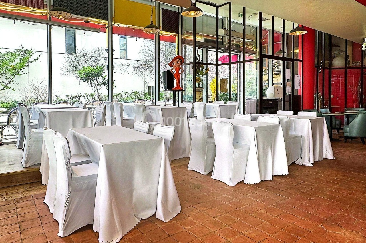 Salle de restaurant lumineuse avec des tables et chaises recouvertes de nappes et housses blanches, vue sur l'extérieur.