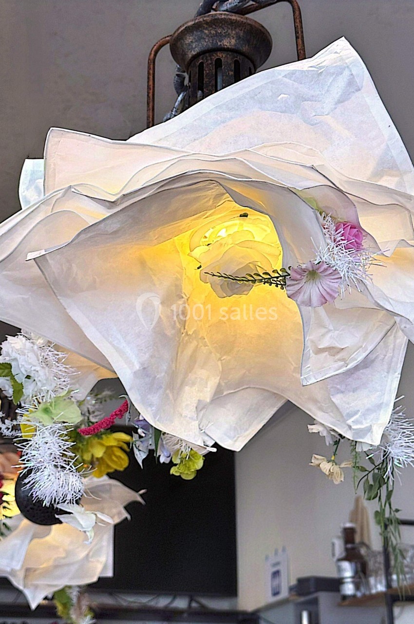 Suspension lumineuse en forme de fleur blanche avec des décorations florales colorées, éclairée dans un intérieur.